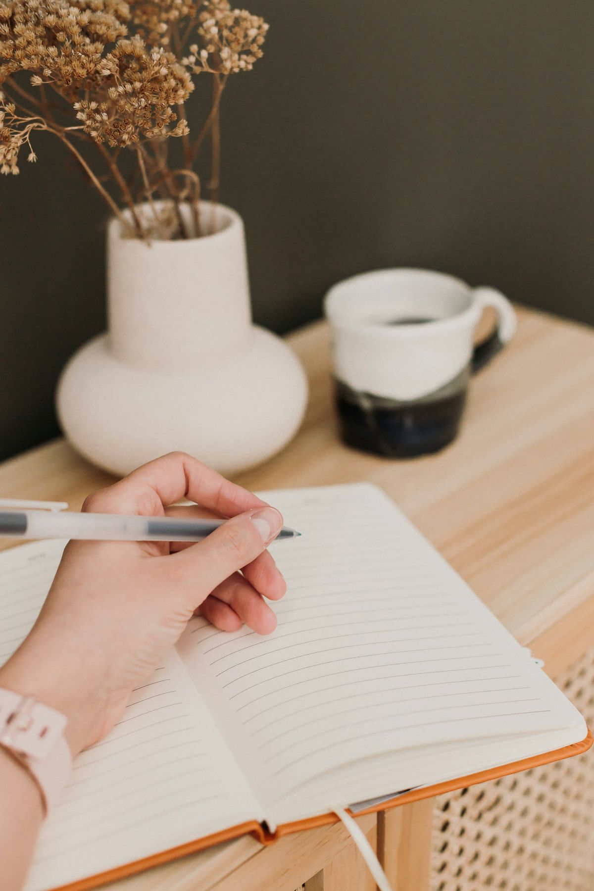 A woman writing in an open journal beside a mug of coffee and dried flowers